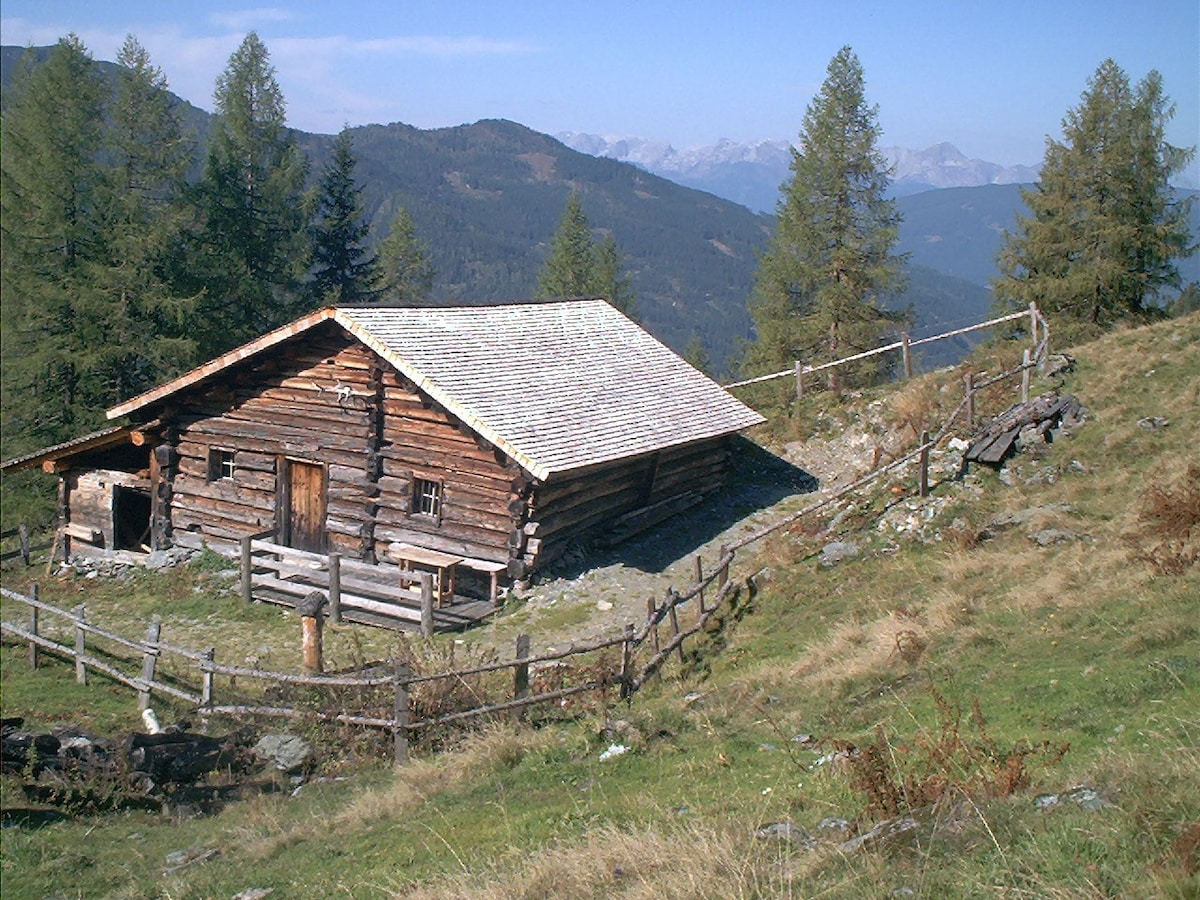 A rustic wooden cabin is situated on a gently sloping hillside, surrounded by trees and grass. The structure features a sloped roof and a wooden door. A simple fence encloses the property, extending towards a nearby path that leads into the natural landscape with distant mountains visible in the background.