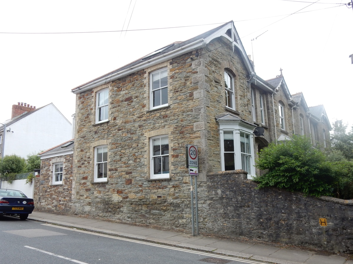 The exterior of a two-story stone house is displayed, featuring large windows that allow natural light to enter. The structure is positioned near the street, with a well-maintained garden area visible beside the building. The surrounding neighborhood appears quiet and residential.