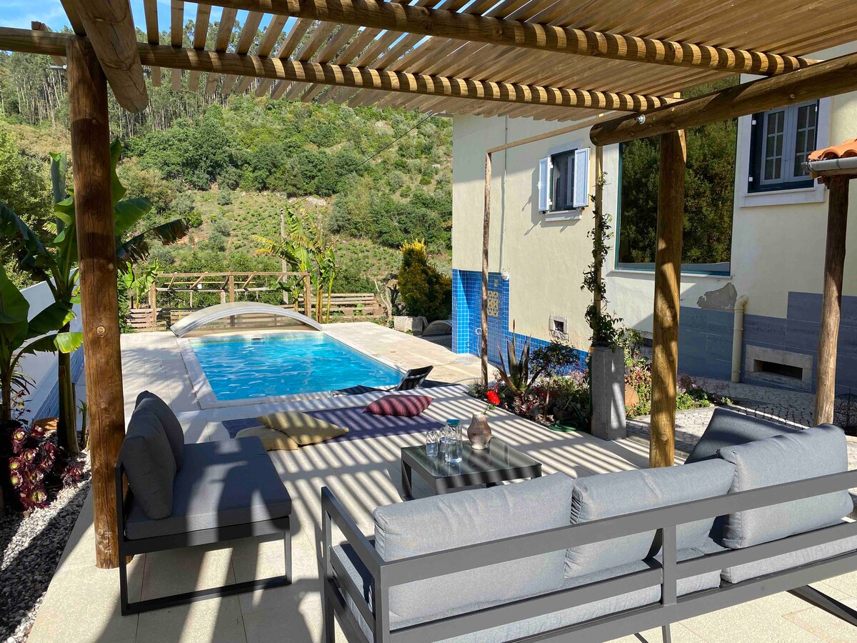 An outdoor seating area is shaded by a wooden pergola, featuring a grey sectional sofa and a coffee table. A private pool is visible in the background, surrounded by lush greenery and stone pathways. Sunlight streams through the slats of the pergola, creating dappled patterns.