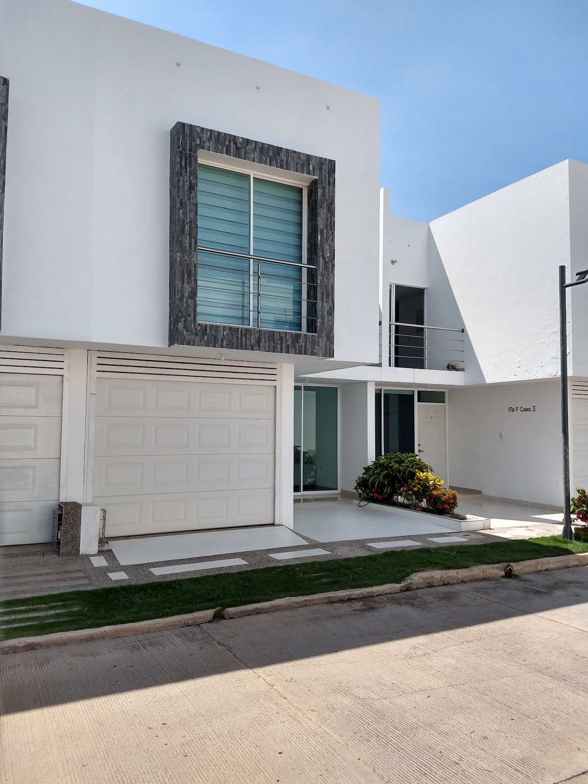 A modern two-story building is presented with a sleek white facade, accented by dark stone frames around large windows. A covered entrance is visible, along with a neatly landscaped area that includes colorful plants and a pathway leading to the door.