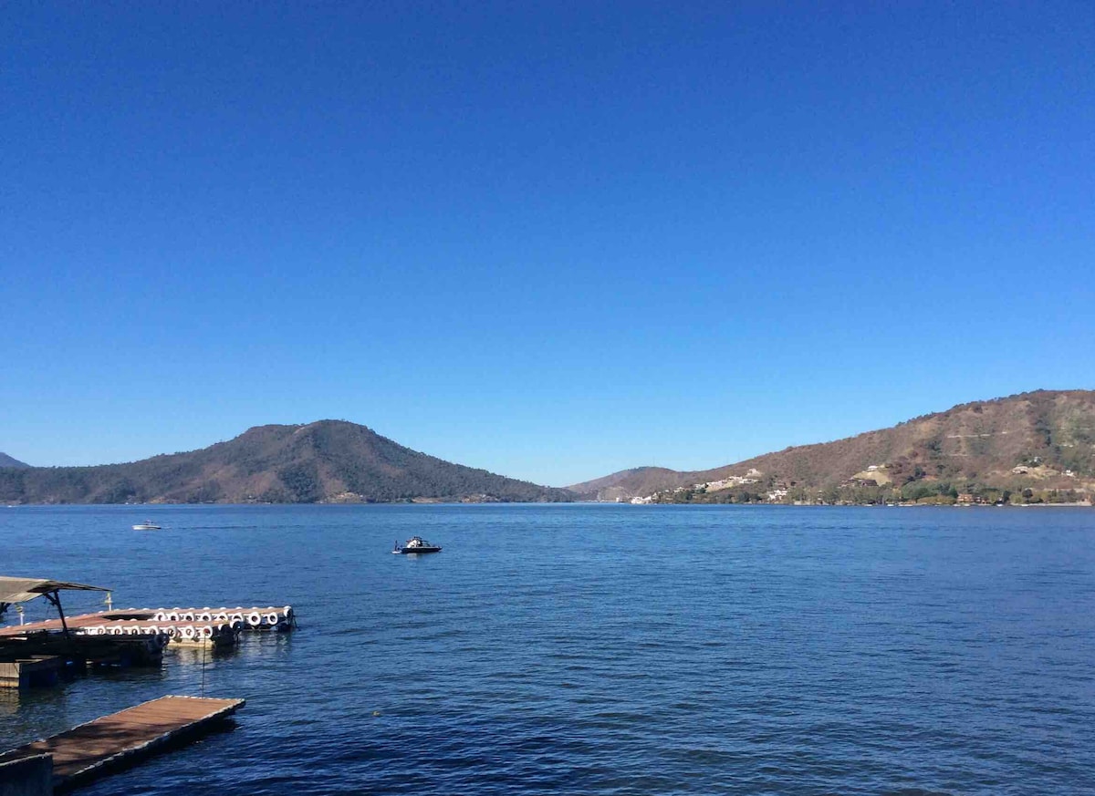 A serene view of a lake bordered by rolling hills under a clear blue sky. The calm water reflects the landscape, with a small boat visible in the distance. Wooden docks are positioned along the shoreline, providing a tranquil setting.