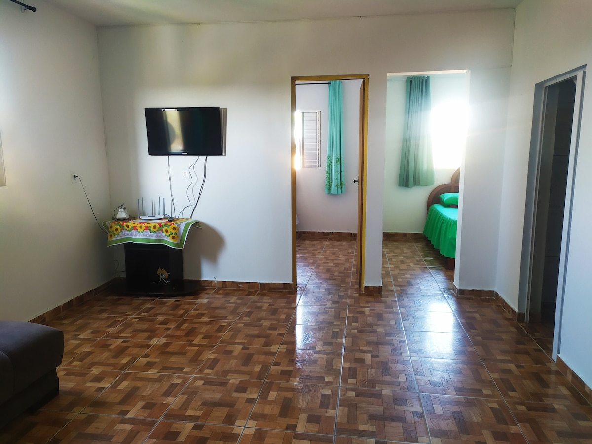 A spacious living area features a wall-mounted television and a comfortable seating arrangement. Light-colored walls enhance the openness. Doorways lead to additional rooms, while large windows allow natural light to brighten the space, complemented by green curtains.