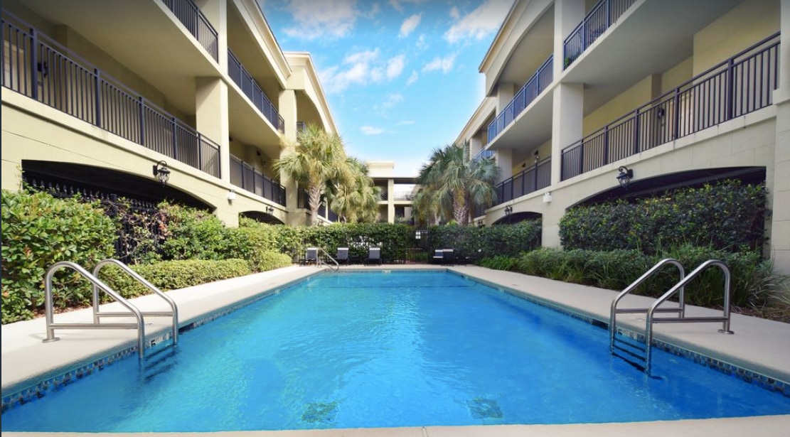 A clear blue pool is framed by lush greenery and palm trees. The surrounding area features two levels of covered balconies from the building, creating a shaded atmosphere. Poolside lounge chairs are visible, inviting relaxation under the sunny sky.