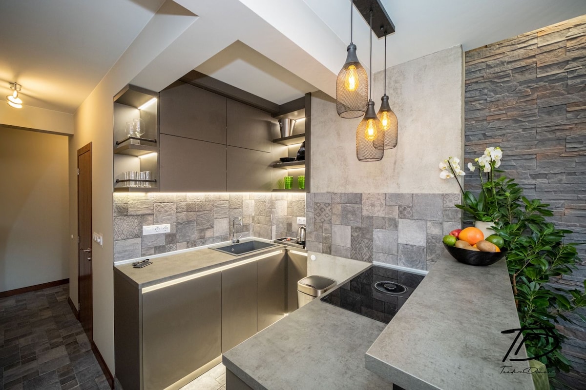 A modern kitchen features sleek cabinetry and an under-cabinet lighting design that highlights the countertop. A small fruit bowl with assorted fruits adds a touch of color. Elegant pendant lights hang above the bar area, while a textured stone wall provides visual interest.