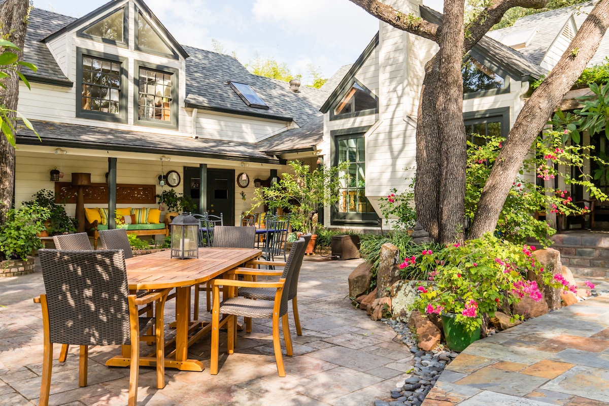 A spacious outdoor patio area is featured, furnished with a wooden dining table surrounded by chairs. Lush greenery and colorful flowers frame the scene, while sunlight filters through the trees, creating a serene atmosphere. The entrance to the guesthouse is visible, blending with the natural surroundings.