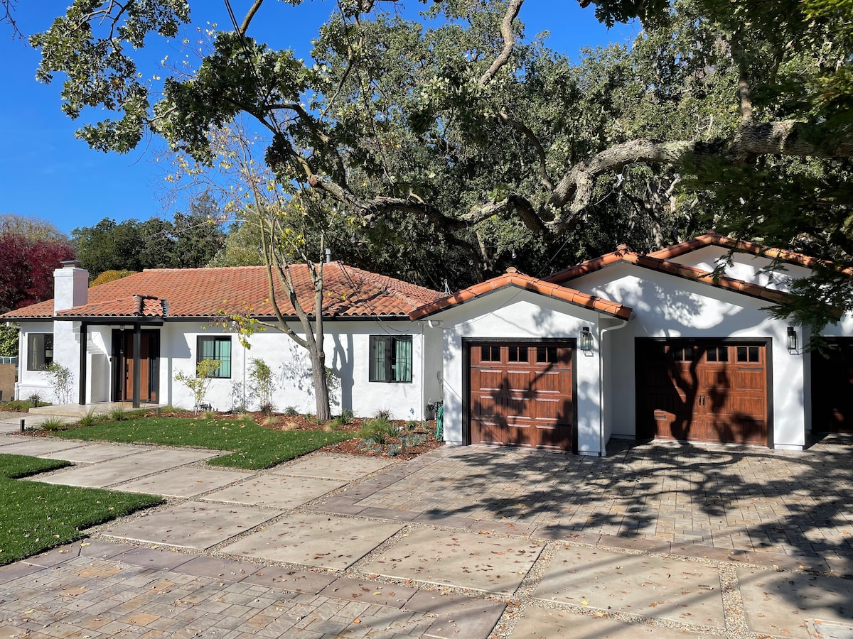 A charming house with terracotta roof tiles is set against a clear blue sky. The front yard features neatly manicured grass, and a paved driveway leads to two wooden garage doors. Trees provide shade, enhancing the welcoming exterior of the home.