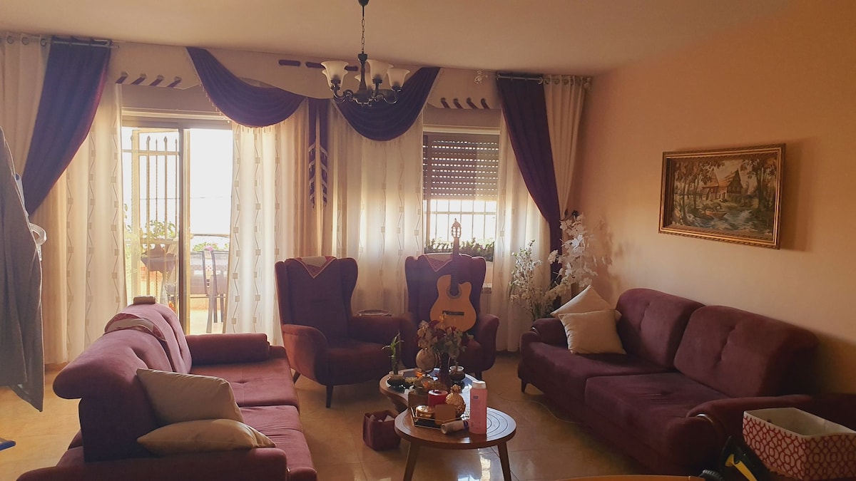 A spacious living area is presented with rich, maroon furniture, including two armchairs and multiple sofas. Natural light filters through sheer curtains, illuminating the room. A round wooden table is situated at the center, adorned with a vase of flowers and various items.