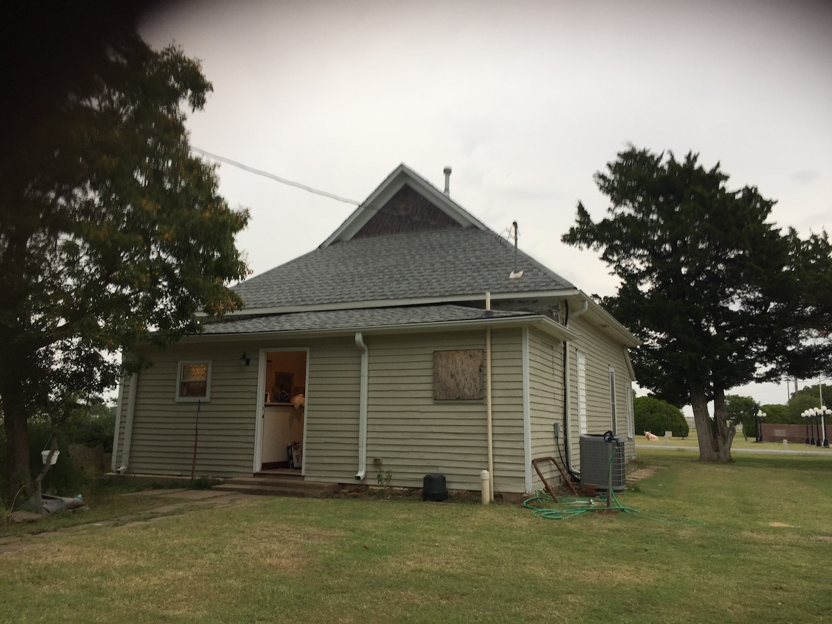 The exterior of a quaint, renovated house features a gray roof and light-colored siding. Windows are visible, with one partially covered. The yard is grassy, bordered by a few large trees. A peaceful setting is created by the surrounding open space.