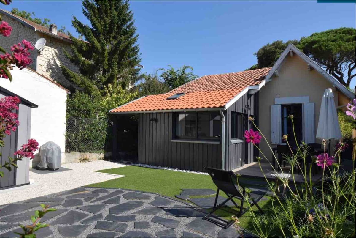 A charming exterior view of a renovated maison is shown, featuring a traditional terracotta roof and grey wooden accents. Surrounding gardens include vibrant flowers, manicured green lawn, and a stone pathway. A sitting area with a black chair and a white parasol invite relaxation.