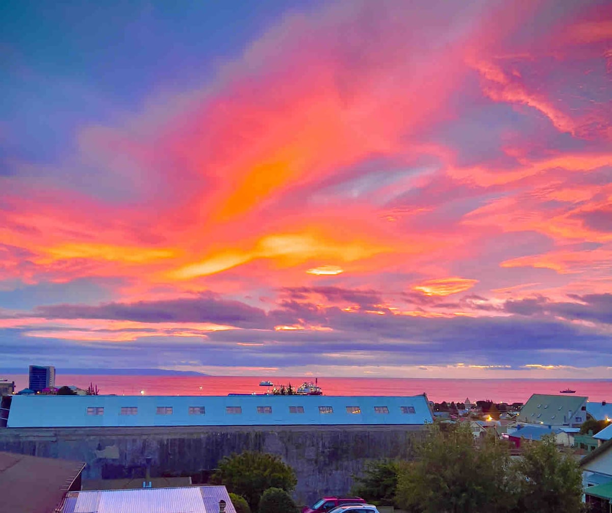 A vibrant sunset paints the sky in hues of orange, pink, and purple above a tranquil ocean view. The silhouette of buildings can be seen in the foreground, with the water reflecting the stunning colors of the evening sky.