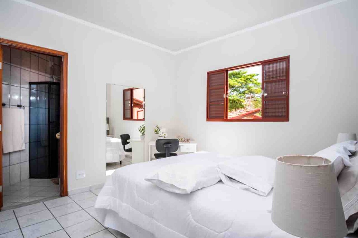 A well-lit bedroom features a spacious bed with white linens, complemented by a chair and desk nearby. A window allows natural light to filter in, revealing views of greenery outside. The ensuite bathroom is visible through an open door.