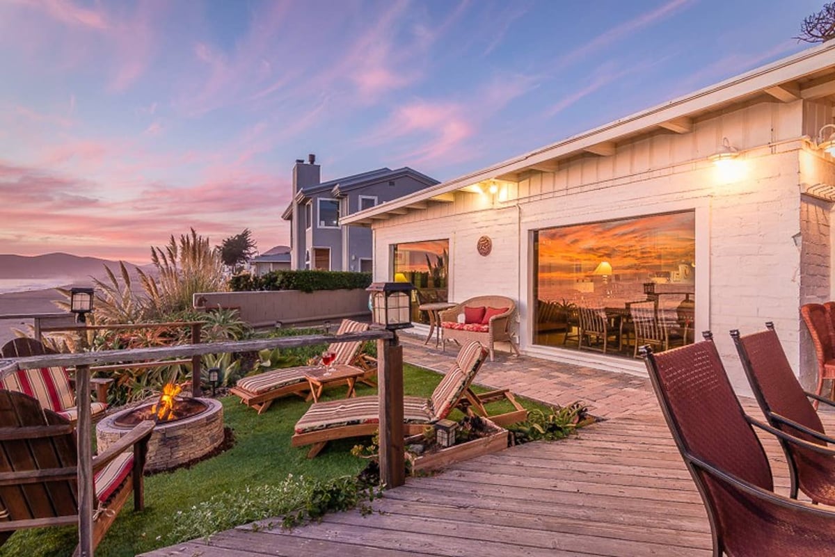 An outdoor seating area is presented with various lounge chairs arranged around a fire pit. Lush greenery and decorative plants enhance the space, while expansive glass doors reflect a sunset view and create a connection to the indoor living area.