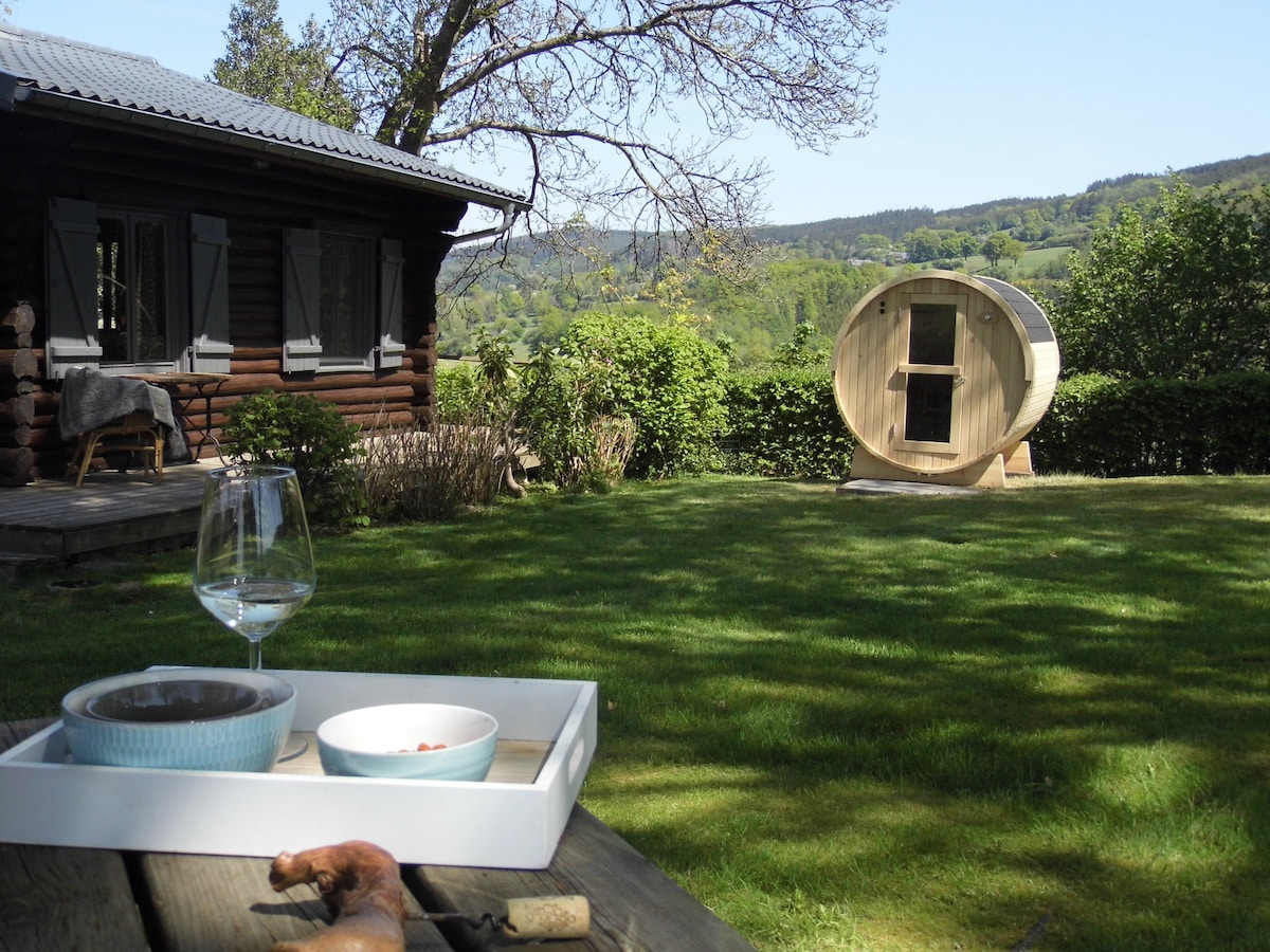 A wooden chalet is visible in the background, surrounded by lush greenery. In the foreground, a glass of wine and a bowl of snacks are arranged on a tray. A unique sauna structure adds interest to the garden area, set against the scenic valley.