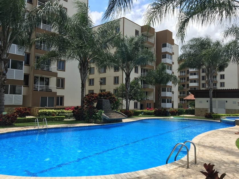 A clear swimming pool is surrounded by palm trees and colorful flower beds within the condominium complex. Modern apartment buildings are positioned in the background, showcasing balconies and lush greenery. Sunlight reflects off the water, creating a serene atmosphere for relaxation.