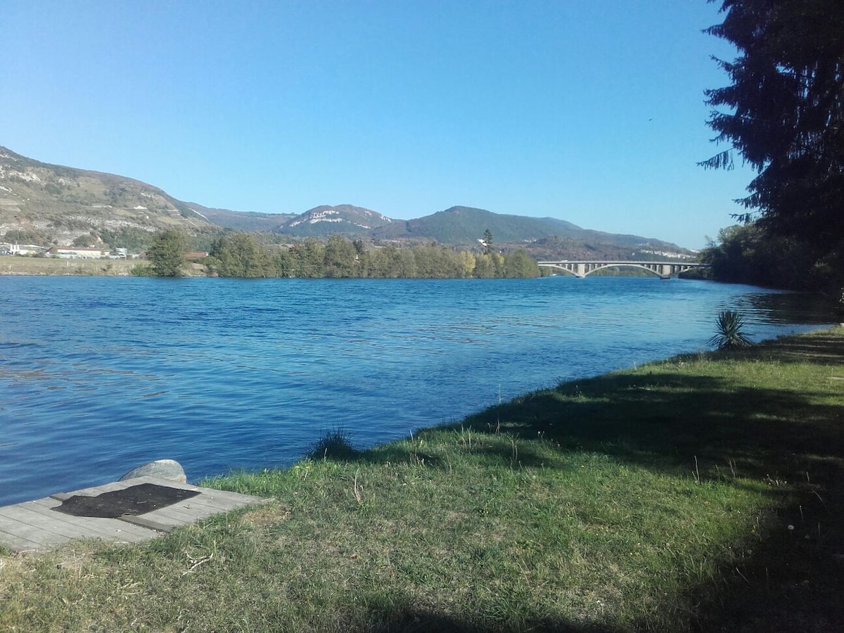 A serene view of a river is captured, showcasing clear blue waters reflecting the sky. Lush greenery lines the riverbank, and distant mountains provide a natural backdrop. A bridge spans the river, creating a connection between the banks.