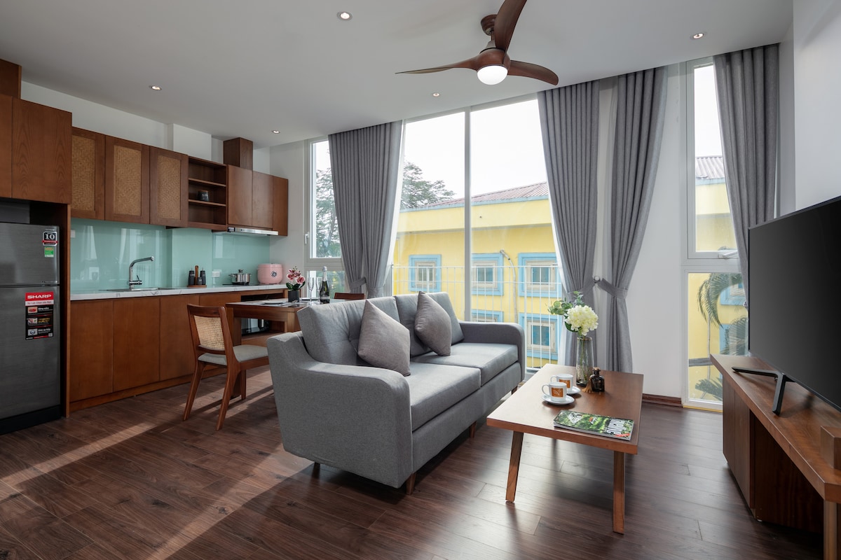 A modern living space features a comfortable grey sofa paired with decorative pillows and a wooden coffee table. Large windows allow natural light to fill the room, while a well-equipped kitchen area is visible in the background. Subtle decor elements enhance the inviting atmosphere.