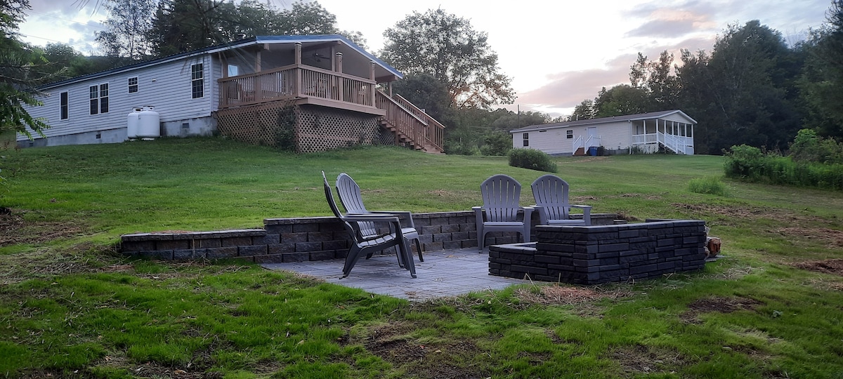 A stone patio is featured in the foreground, equipped with several gathered chairs surrounding a fire pit. The background displays a well-maintained home, set on a grassy lawn and framed by trees, with a large covered deck accessible from the side.