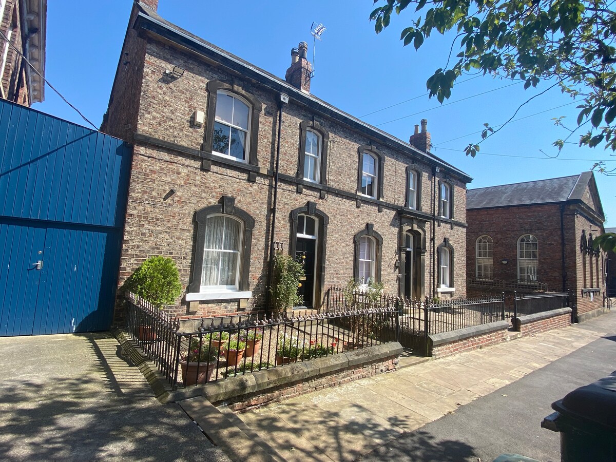 The exterior of a charming brick building is shown, featuring a well-maintained garden with greenery and flower beds. A gated entrance leads to the property, which showcases multiple windows aligned symmetrically. Soft sunlight brightens the facade, enhancing the inviting appearance of the home.