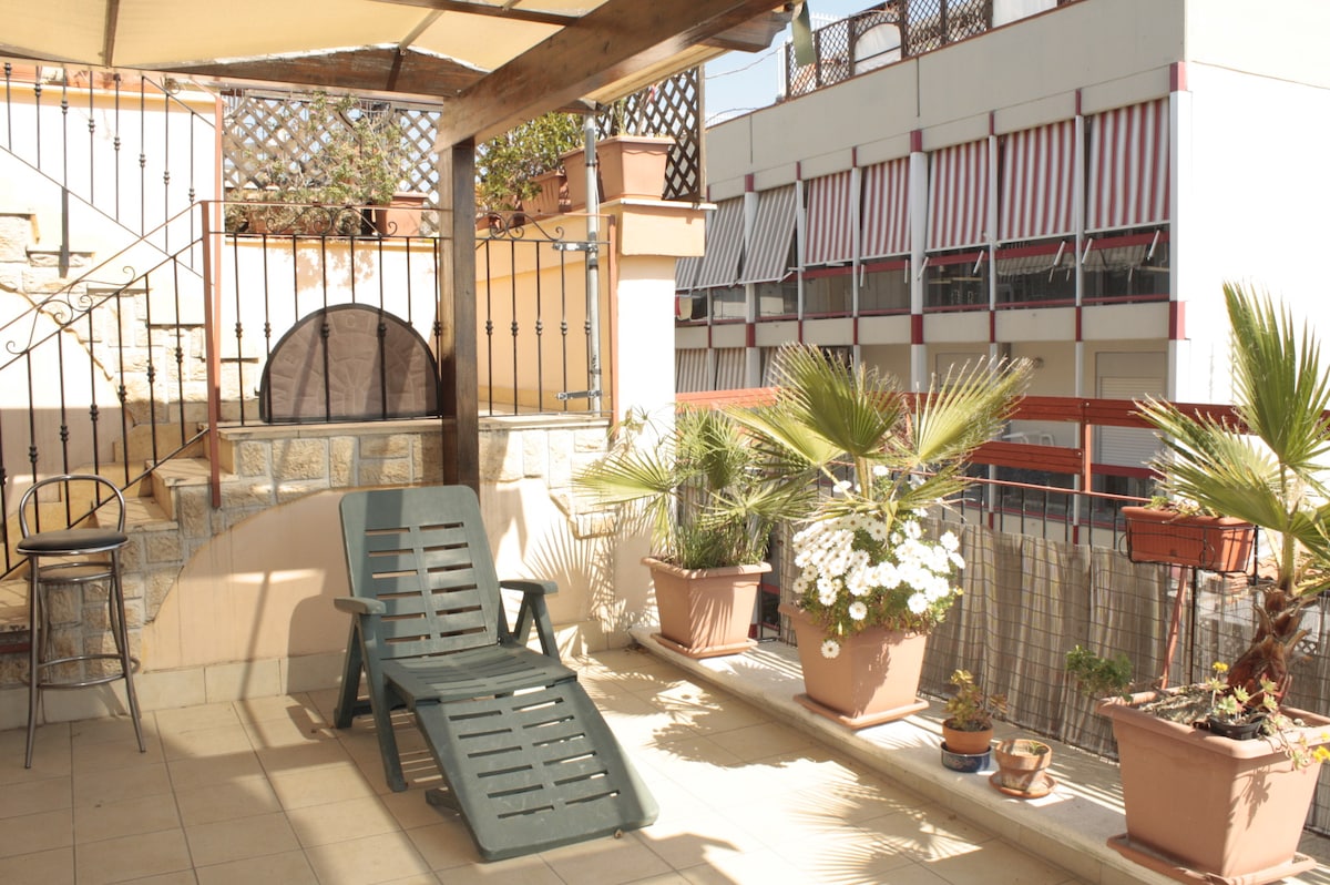 A terrace space is featured, furnished with a reclining lounge chair and surrounded by various potted plants. The area is bathed in natural light, with a sunshade providing comfort. The backdrop includes a glimpse of neighboring buildings, enhancing the urban setting.