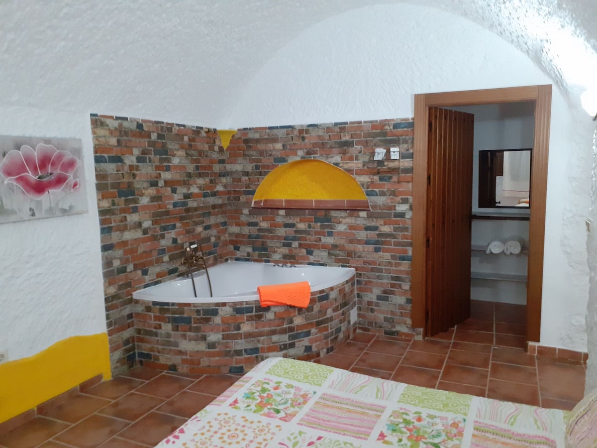 The room features an inviting jacuzzi, surrounded by textured brick walls. A colorful towel is draped over the side. A doorway leads to an adjoining bathroom area, while a bed with a floral patterned blanket is positioned in the foreground.