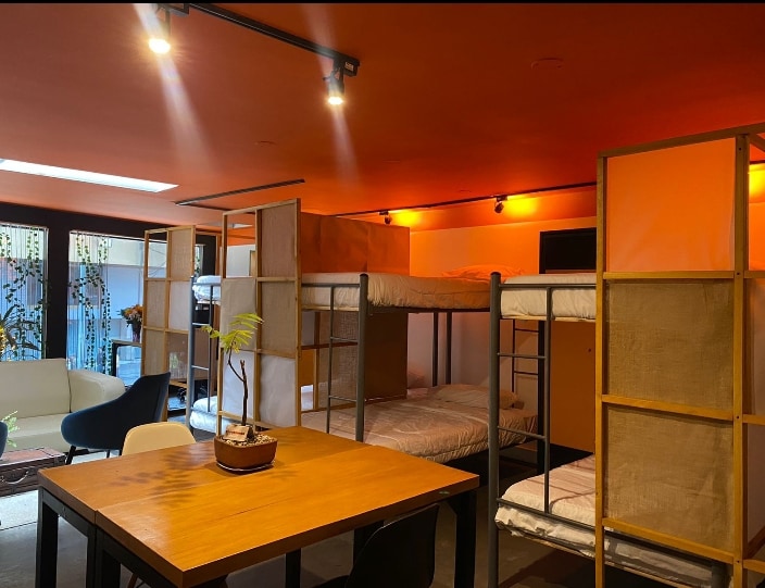 A communal sleeping area features multiple bunk beds with simple frameworks, allowing for shared accommodations. Soft orange lighting creates a warm ambiance, while a wooden table and chairs are situated nearby, promoting a welcoming environment for interactions.