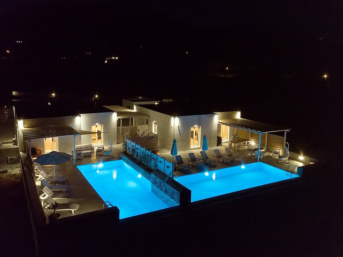 Two separate villas feature illuminated private swimming pools, exuding a calming blue glow against the night sky. Sun loungers and umbrellas are arranged around the pools, enhancing outdoor relaxation. The surrounding area is tranquil, with distant lights subtly visible in the background.