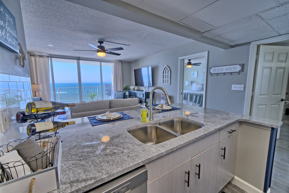 An updated kitchen is visible, featuring granite countertops and stainless steel appliances. A wide open layout leads to a spacious living area, with floor-to-ceiling windows offering views of the Gulf of Mexico. A ceiling fan provides airflow in the inviting space.