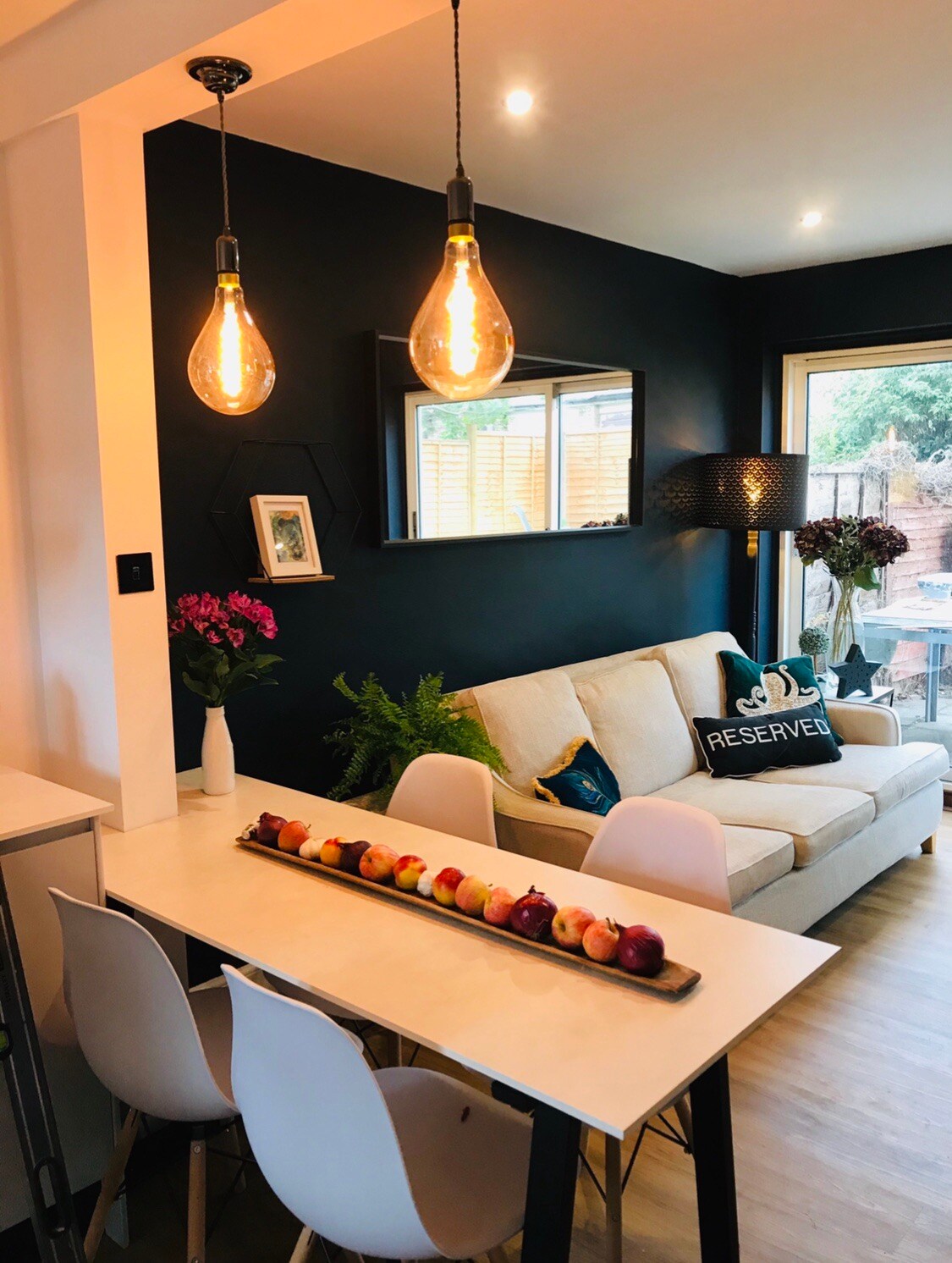 A modern dining area is featured, showcasing a long wooden table adorned with a selection of colorful apples. White chairs surround the table, while a cozy sofa is situated nearby. Soft lighting is provided by hanging bulbs, illuminating the dark blue walls and a mirror.
