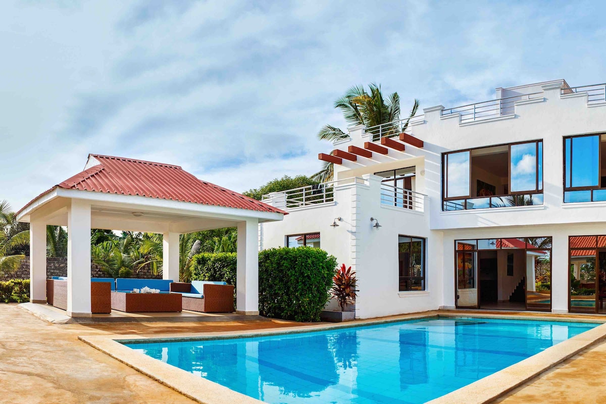 A modern villa features a sparkling blue pool adjacent to a shaded seating area with a thatched roof. Lush green plants surround the property, and large windows reflect a clear sky, enhancing the airy atmosphere of the outdoor space.