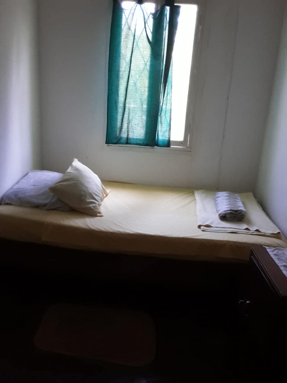 A simple sleeping area features a twin bed dressed in a light yellow coverlet. Two pillows sit at the head of the bed, with a rolled towel placed neatly beside it. A window with teal curtains allows natural light to filter into the space.