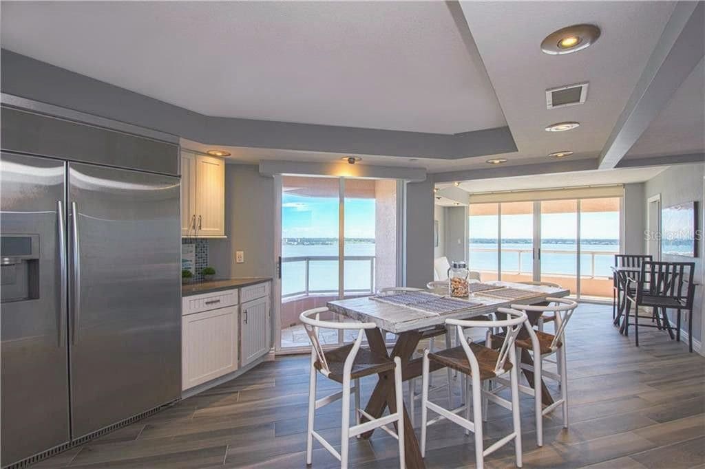 An open kitchen area features modern stainless-steel appliances, white cabinetry, and a spacious island with seating. Large glass doors allow natural light and offer views of the water, with a dining area positioned beside the windows, creating a seamless connection to the outdoor scenery.