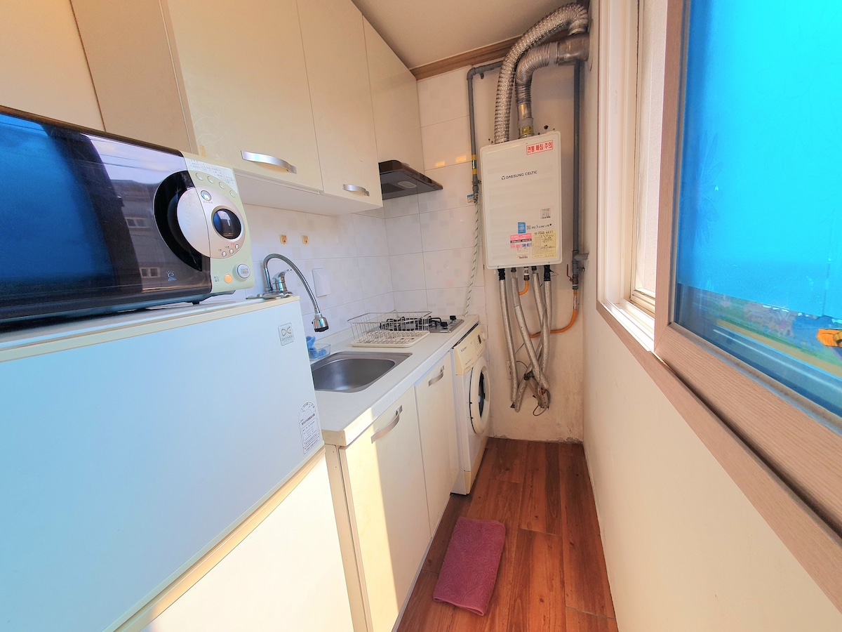A compact kitchen area is equipped with a sink, stove, and refrigerator. A washing machine is also present. Light wood flooring complements the modern cabinetry. A window offers a view, enhancing the space with natural light.