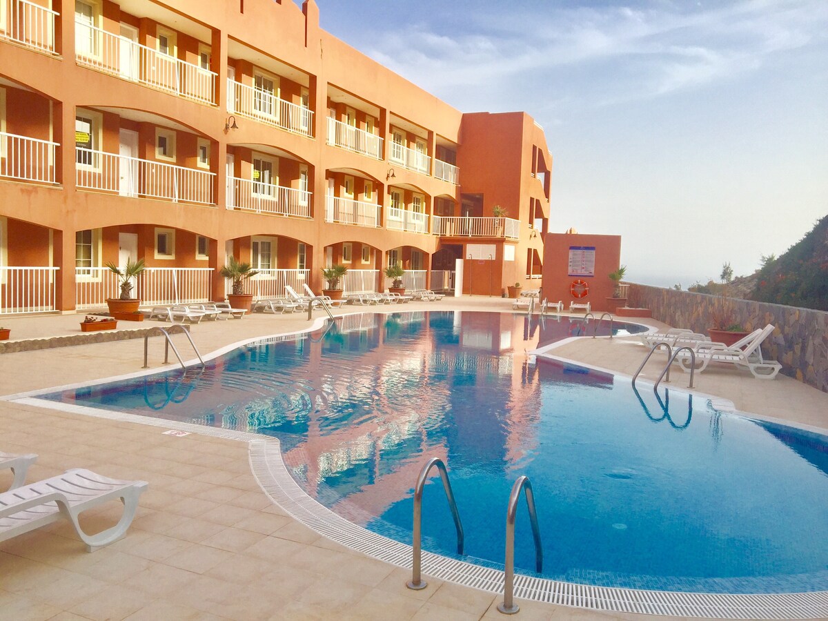 A large outdoor swimming pool is surrounded by sun loungers and palm plants, reflecting the soft colors of the building façade in the background. The pool area is well-lit under a clear sky, providing a serene atmosphere for relaxation.