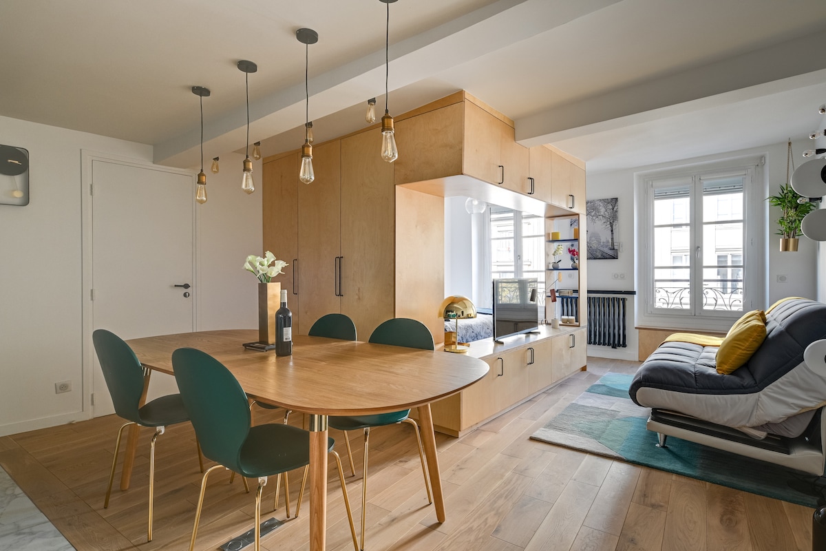 A modern dining area features a round wooden table surrounded by green upholstered chairs. Above, stylish pendant lights hang from the ceiling. A sleek wall unit holds decorative items, while a comfortable sofa in the corner offers a cozy sitting space next to a window with natural light.