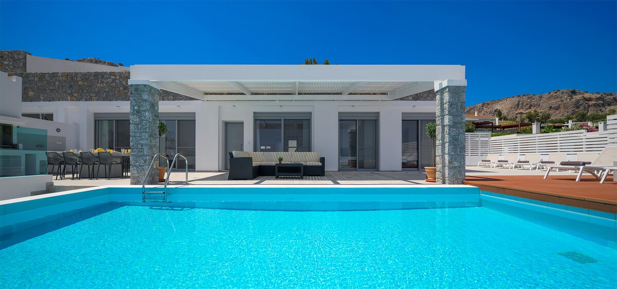 A modern outdoor space is featured, showcasing a swimming pool with clear blue water. A covered terrace area is visible, furnished with seating. Surrounding elements include neatly arranged lounge chairs and a scenic backdrop of gentle hills under a bright blue sky.