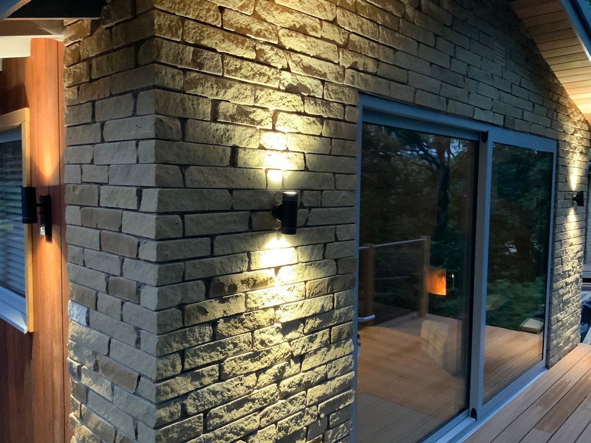An exterior wall features a textured stone finish, enhancing the chalet's rustic character. Soft lighting is provided by fixtures mounted alongside the large glass doors, which reflect the surrounding greenery. A wooden deck is visible, complementing the natural materials used in construction.