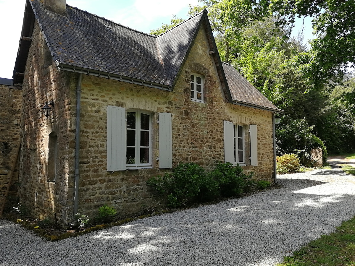 A rustic stone cottage is nestled among lush greenery, with soft gravel leading to its entrance. White shutters frame the windows, which reflect natural light. The peaked roof adds character, blending harmoniously with the surrounding trees that enhance the peaceful environment.