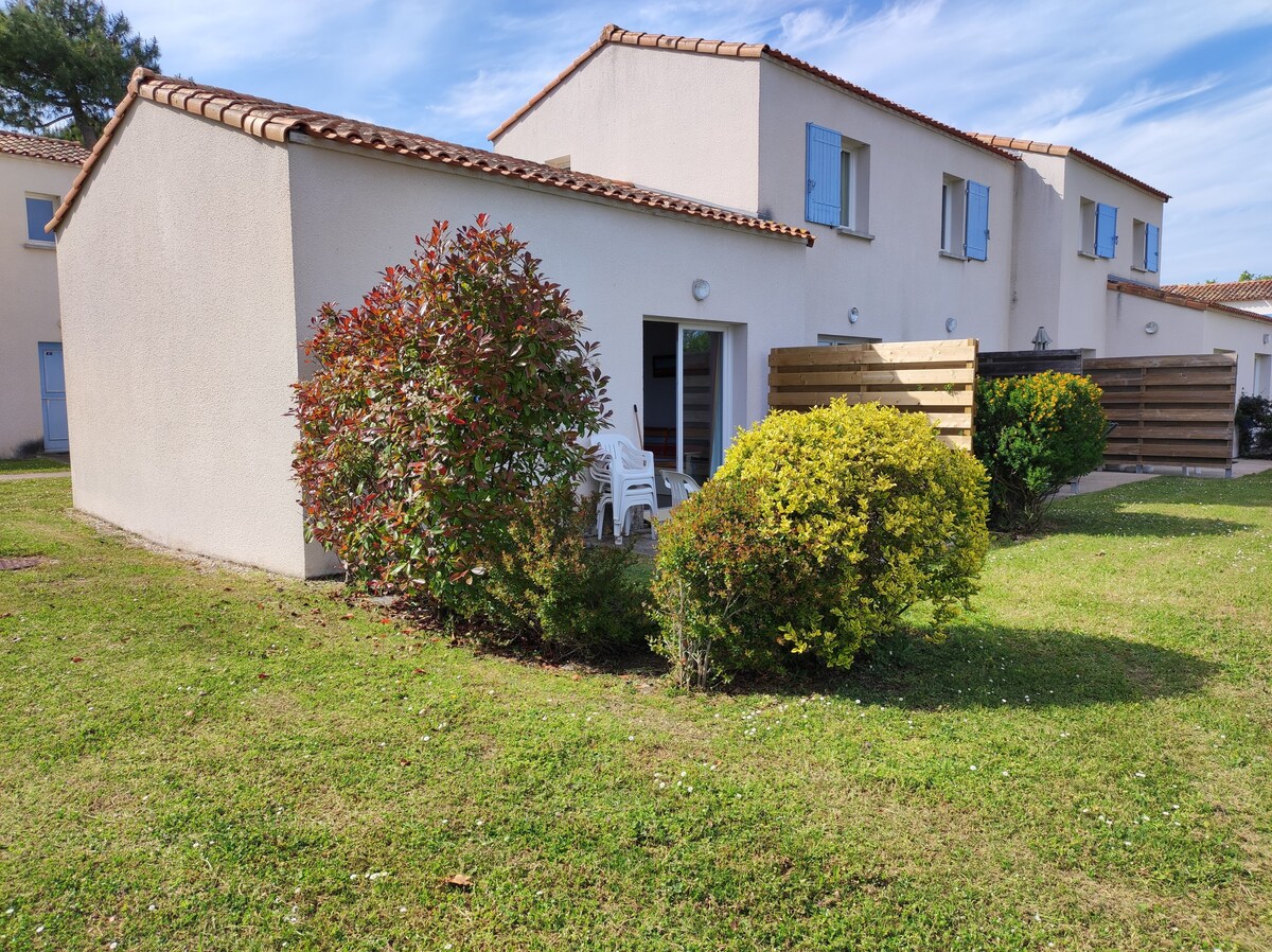 A single-story building is surrounded by well-maintained greenery, featuring a small patio with a white chair visible at the entrance. The exterior walls are light-colored, and blue shutters decorate the windows, giving a serene appearance to the space.