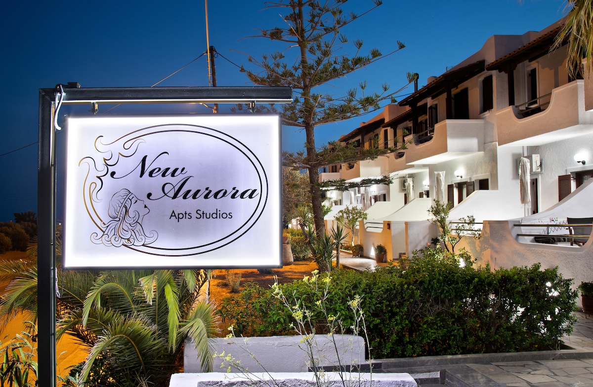 The entrance sign of New Aurora Apts Studios is illuminated, welcoming guests to the complex. The building features a modern design with white exterior walls and multiple balconies. Lush greenery is visible in the foreground, adding a touch of nature to the setting.