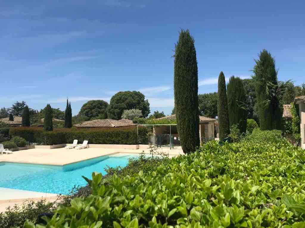A serene outdoor area features a crystal-clear swimming pool surrounded by landscaped greenery. Tall cypress trees and lush hedges provide privacy and enhance the tranquil atmosphere, with terracotta-roofed structures visible in the background under a clear blue sky.