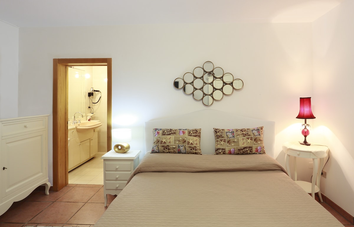 The bedroom features a spacious bed with decorative pillows, complemented by a bedside table with a lamp. A mirror arrangement enhances the room's depth, while a doorway reveals a bathroom area illuminated by warm lighting.