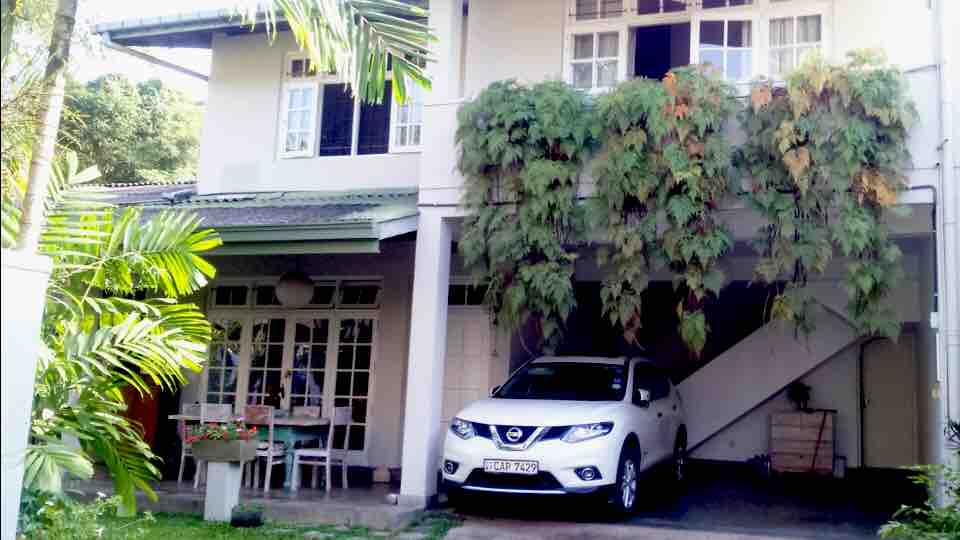 A two-story house features a covered parking area with a white SUV parked beneath it. Lush green foliage trails down from the upper balcony, adding a touch of nature to the structure. A small table and chairs are placed outside under the shade of trees.