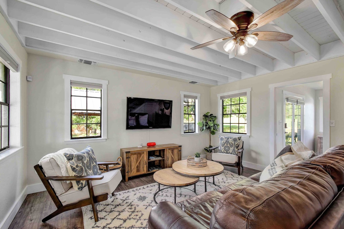 The living area features a smooth wooden floor, a leather sofa, and two armchairs flanking a coffee table. A mounted television is positioned on the wall, while large windows allow natural light to fill the space. Ceiling beams add a touch of architectural interest.