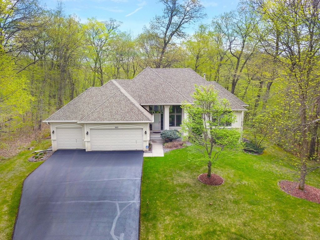 The home is positioned on a spacious lot surrounded by vibrant greenery. A clean driveway leads to a welcoming entrance. The exterior showcases a harmonious blend of soft-toned siding and a welcoming porch area, complemented by manicured landscaping and mature trees.