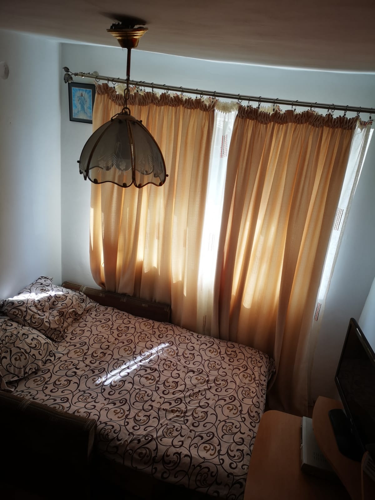 A well-lit sleeping area features a spacious bed adorned with a patterned comforter. Soft, golden curtains filter natural light, while a vintage pendant lamp hangs above. A small television is positioned on a wooden surface, contributing to a functional yet cozy ambiance.