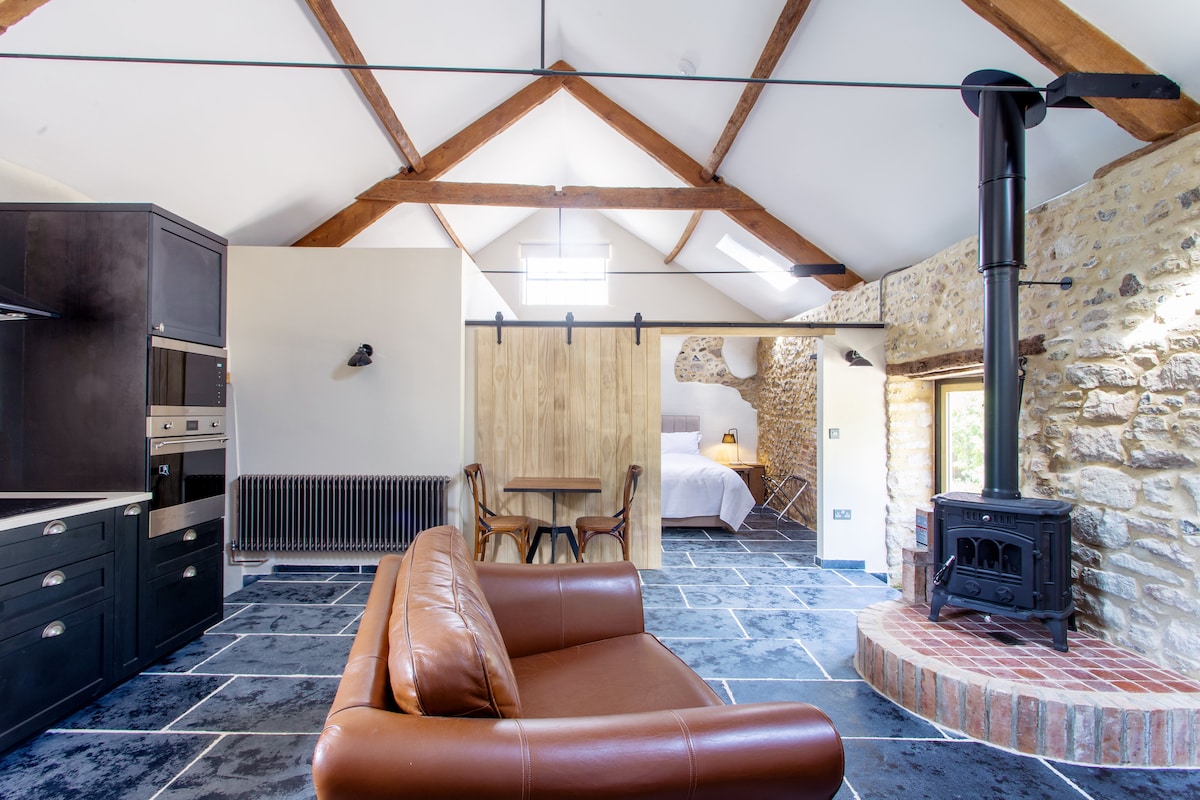 An open living space features a warm color palette with stone walls and wooden beams. A leather sofa is positioned in front of a wood-burning stove, enhancing comfort. A dining area with two chairs is visible, as well as a well-equipped kitchen with modern appliances.