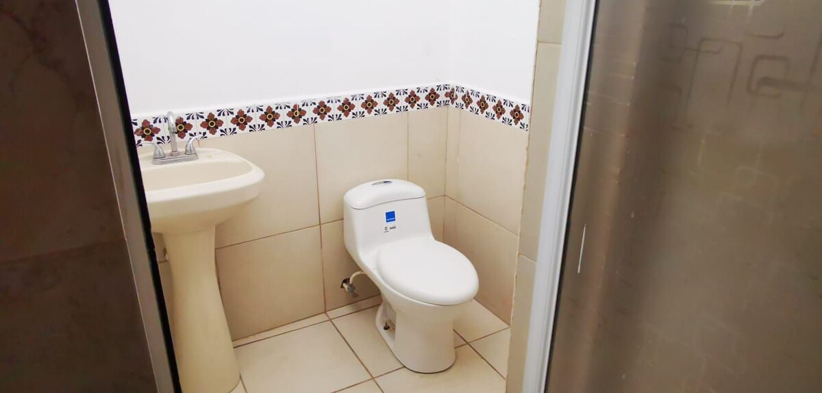 The bathroom features tiled walls with decorative border accents. A modern toilet and pedestal sink are positioned adjacent to one another in a compact layout. Neutral tiles cover the floor, and a transparent shower enclosure is visible on one side.