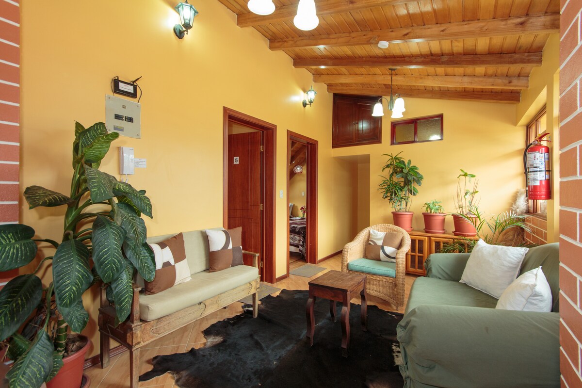 A welcoming communal area is presented with natural wood beams and warm yellow walls. Two seating areas feature a mix of light-colored and dark green sofas. Potted plants add a touch of greenery to the space, while a small wooden table sits centrally amidst the seating.