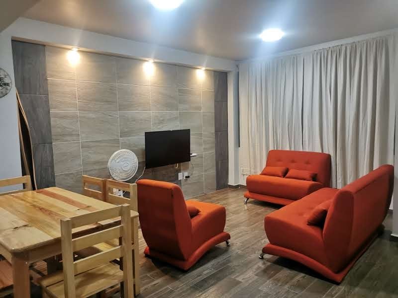 A modern living area features two bright orange sofas, a wooden dining table with chairs, and a flat-screen TV mounted on a textured wall. Soft lighting casts a warm glow, while sheer white curtains frame a window, allowing natural light to fill the space.