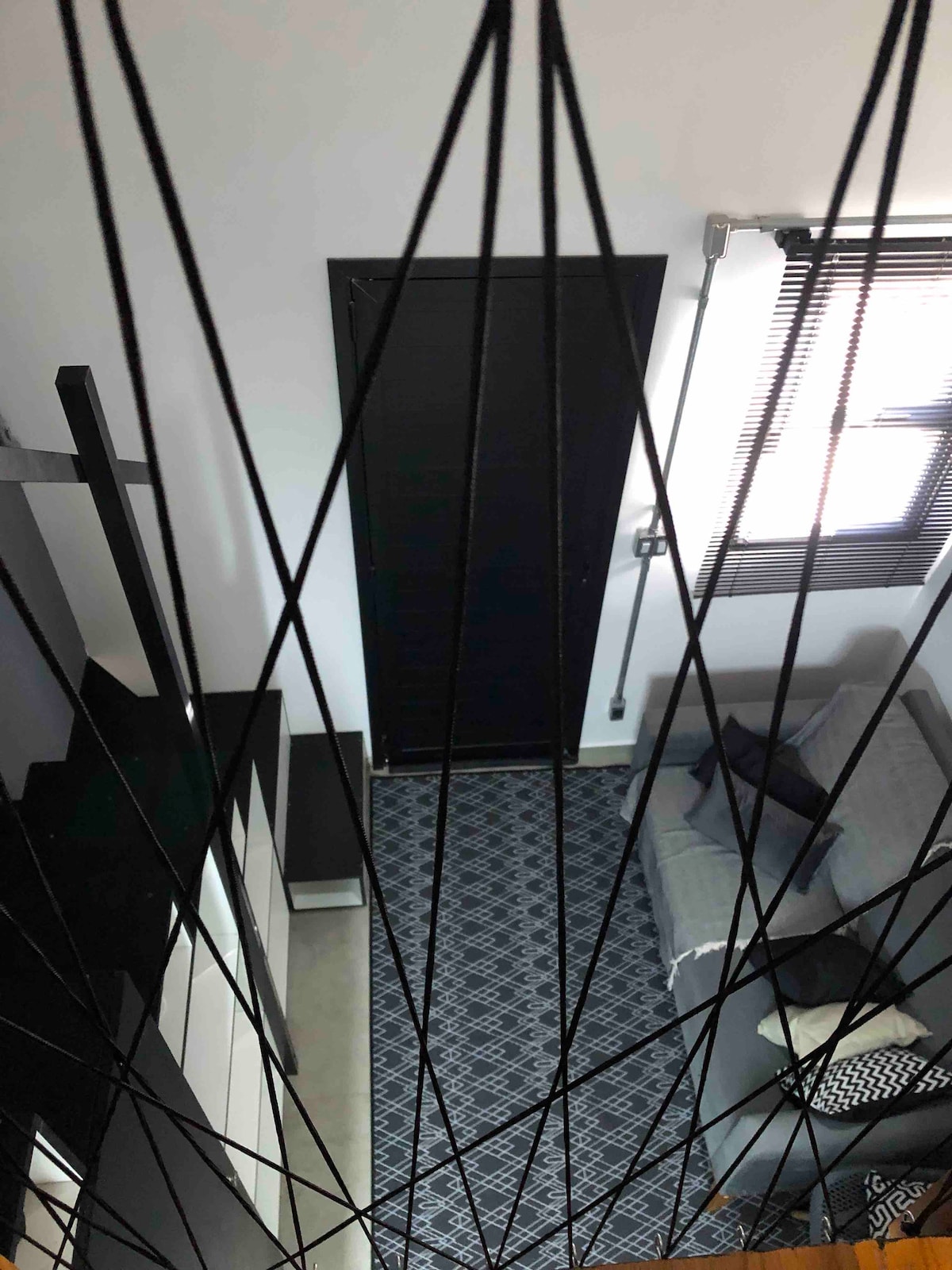 A view from above captures the spacious living area, highlighted by a patterned rug beneath a sofa. A sleek black door is visible, complemented by the geometric lines of ceiling cables that create a modern architectural aspect. Natural light streams in through a blind-covered window.
